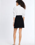 A woman is shown from behind wearing a white long-sleeve button-up shirt and a black mini skirt, paired with black flats, against a plain white studio background.