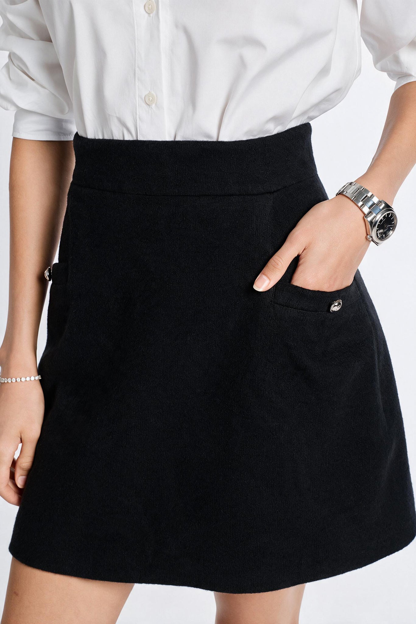 A close-up of a person wearing a black A-line cashmere mini skirt with a front pocket and silver-tone button detail, paired with a white button-up shirt. The person is wearing a silver watch on one wrist and a bracelet on the other, set against a plain white background.