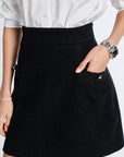 A close-up of a person wearing a black A-line cashmere mini skirt with a front pocket and silver-tone button detail, paired with a white button-up shirt. The person is wearing a silver watch on one wrist and a bracelet on the other, set against a plain white background.