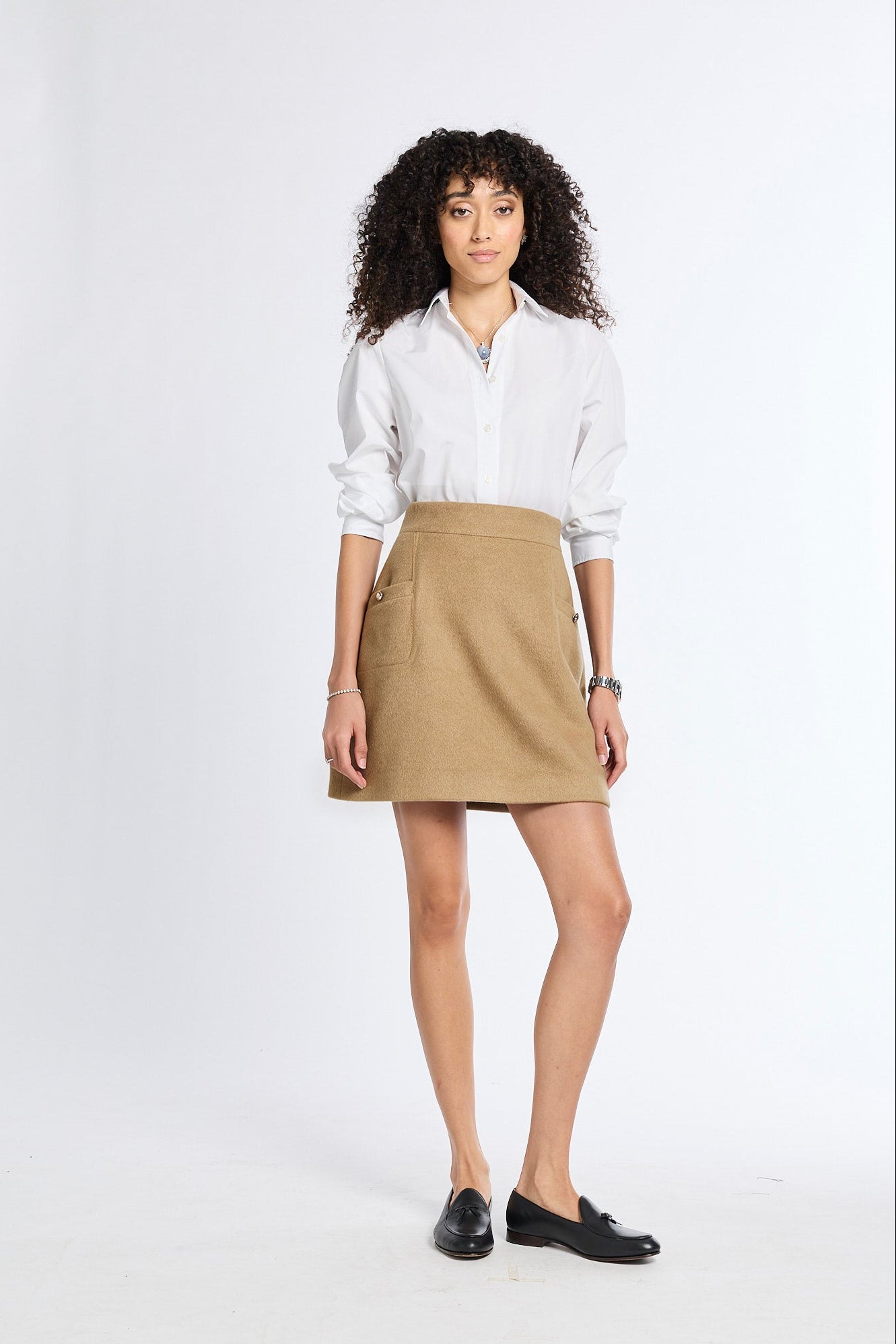 A woman with curly dark hair stands against a white studio background wearing a white button-up shirt, a tan mini skirt, and black loafers.