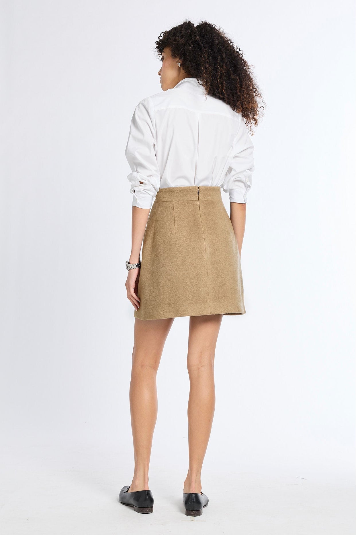 A woman is shown from behind wearing a white long-sleeve button-up shirt and a beige mini skirt, paired with black flats, against a plain white studio background.