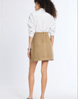 A woman is shown from behind wearing a white long-sleeve button-up shirt and a beige mini skirt, paired with black flats, against a plain white studio background.