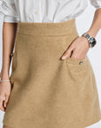 A close-up of a person wearing a tan wool-blend mini skirt with a front pocket, paired with a white button-up shirt. The person is wearing a silver watch on one wrist and a pearl bracelet on the other.