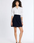 A woman modeling a white long-sleeve button-up shirt and a dark navy blue mini skirt with front patch pockets, paired with black loafers, against a plain white studio background.