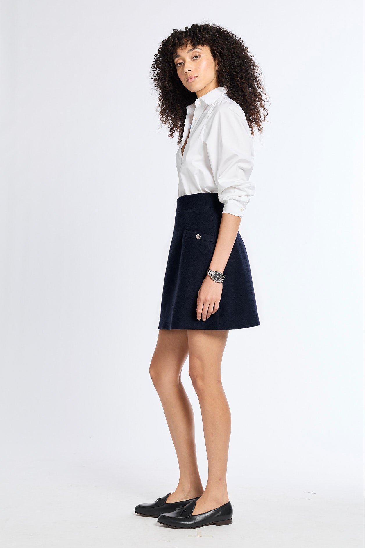 A woman with curly dark hair stands in a studio wearing a white button-up shirt, a dark navy A-line skirt, and black flats against a plain light background.