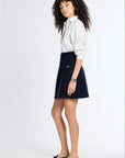 A woman with curly dark hair stands in a studio wearing a white button-up shirt, a dark navy A-line skirt, and black flats against a plain light background.