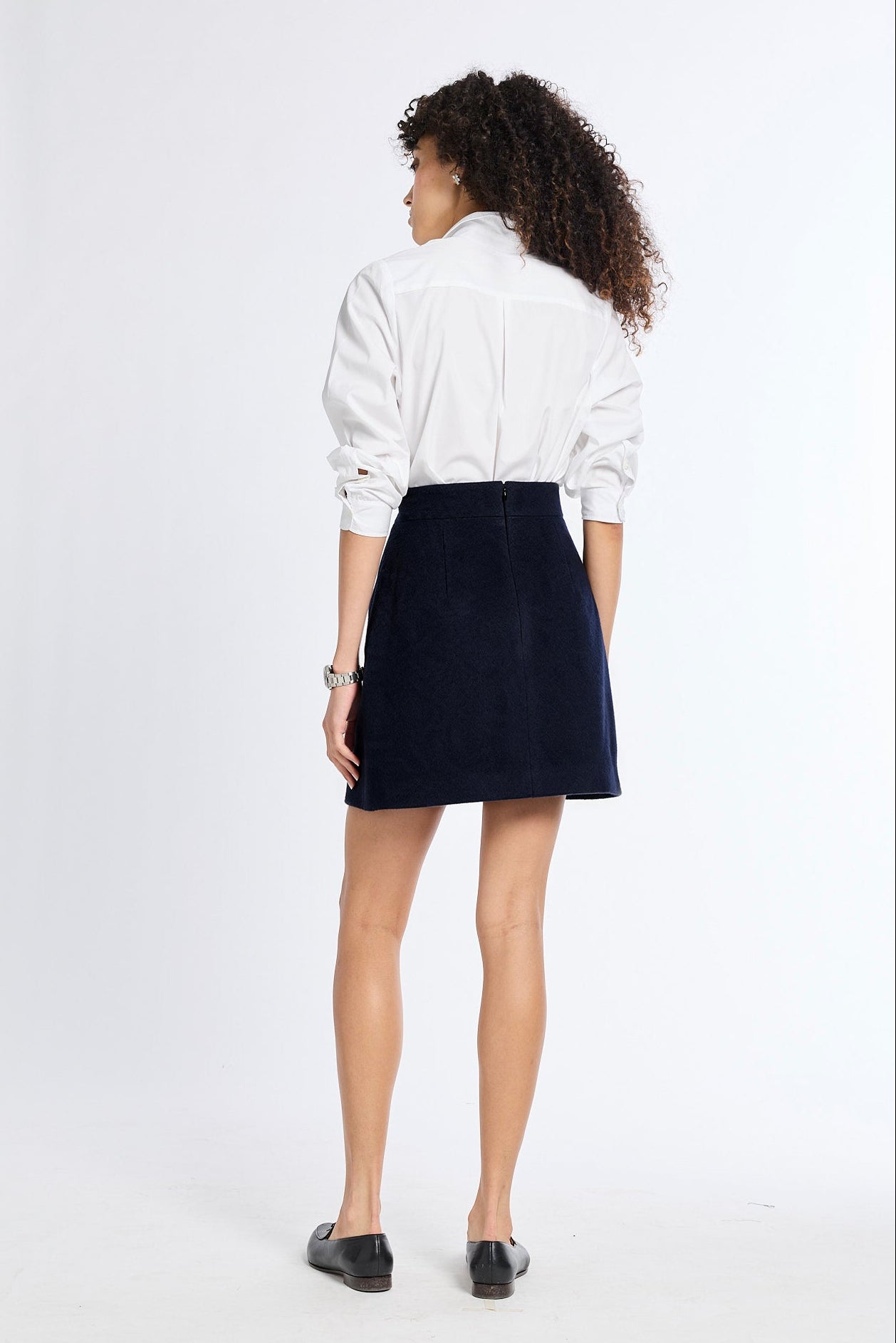 A woman is shown from behind wearing a white long-sleeve button-up shirt and a dark navy blue corduroy mini skirt, paired with black flat shoes, against a plain white studio background.