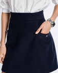 A close-up of a person wearing a dark navy blue A-line skirt with a front pocket, paired with a white button-down shirt. The person is wearing a silver watch on one wrist and a pearl bracelet on the other.