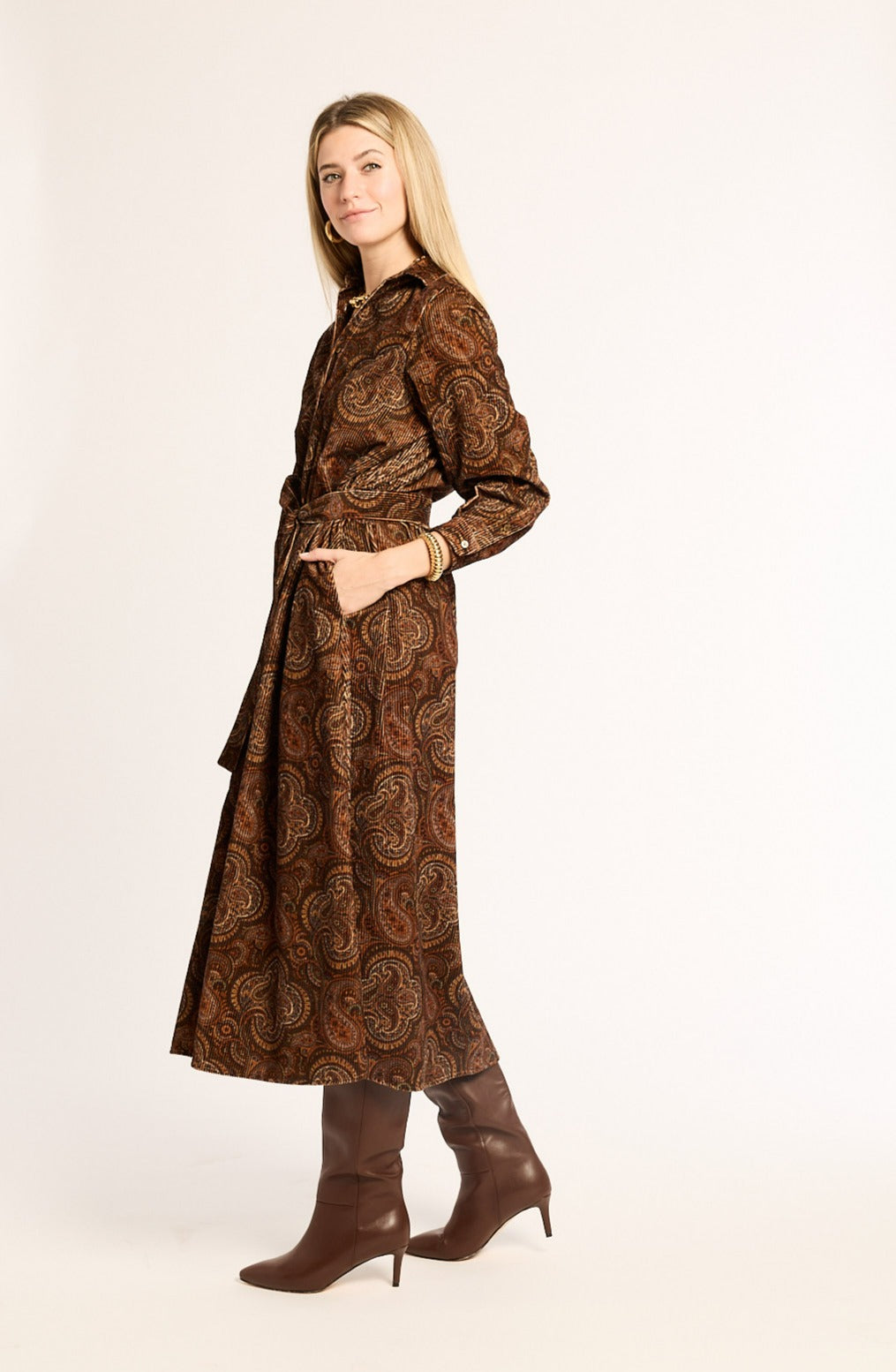 Woman wearing a long brown patterned dress with matching boots on a white background