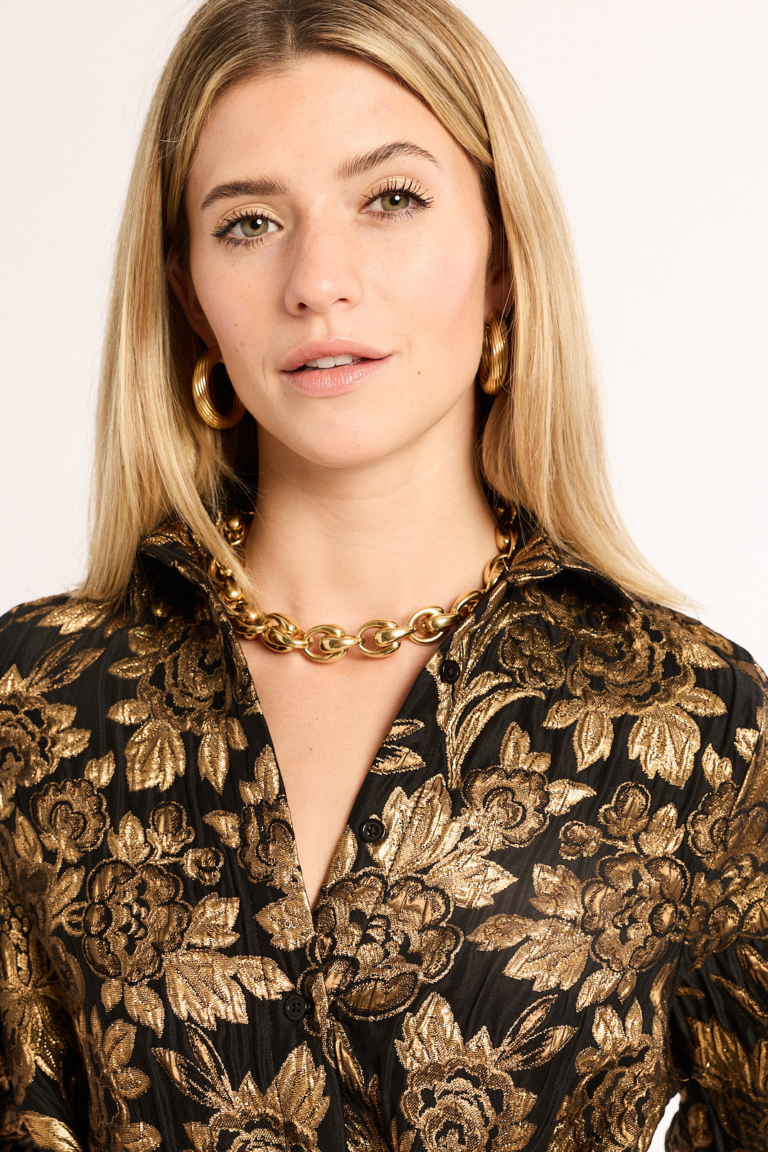 Woman wearing a black and gold floral patterned blouse with a gold necklace against a plain background