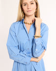 Woman wearing a light blue dress with gold jewelry on a plain background
