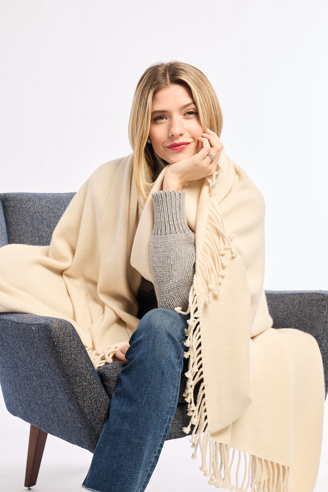 Woman sitting on a gray chair wearing a beige blanket and gray sweater against a white background