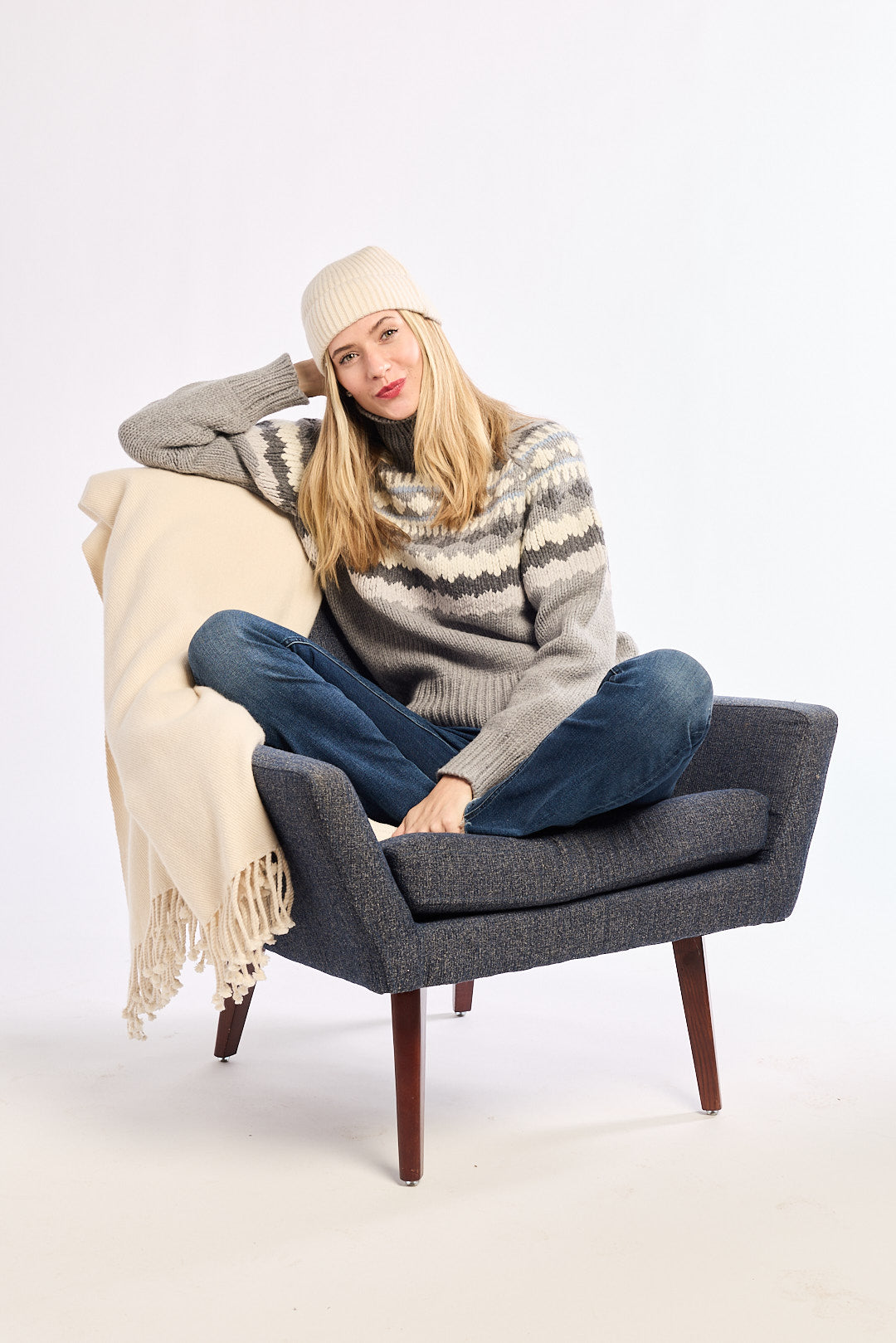Woman sitting on a gray chair with a beige blanket, wearing a patterned sweater and jeans.