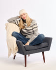 Woman sitting on a gray chair with a beige blanket, wearing a patterned sweater and jeans.