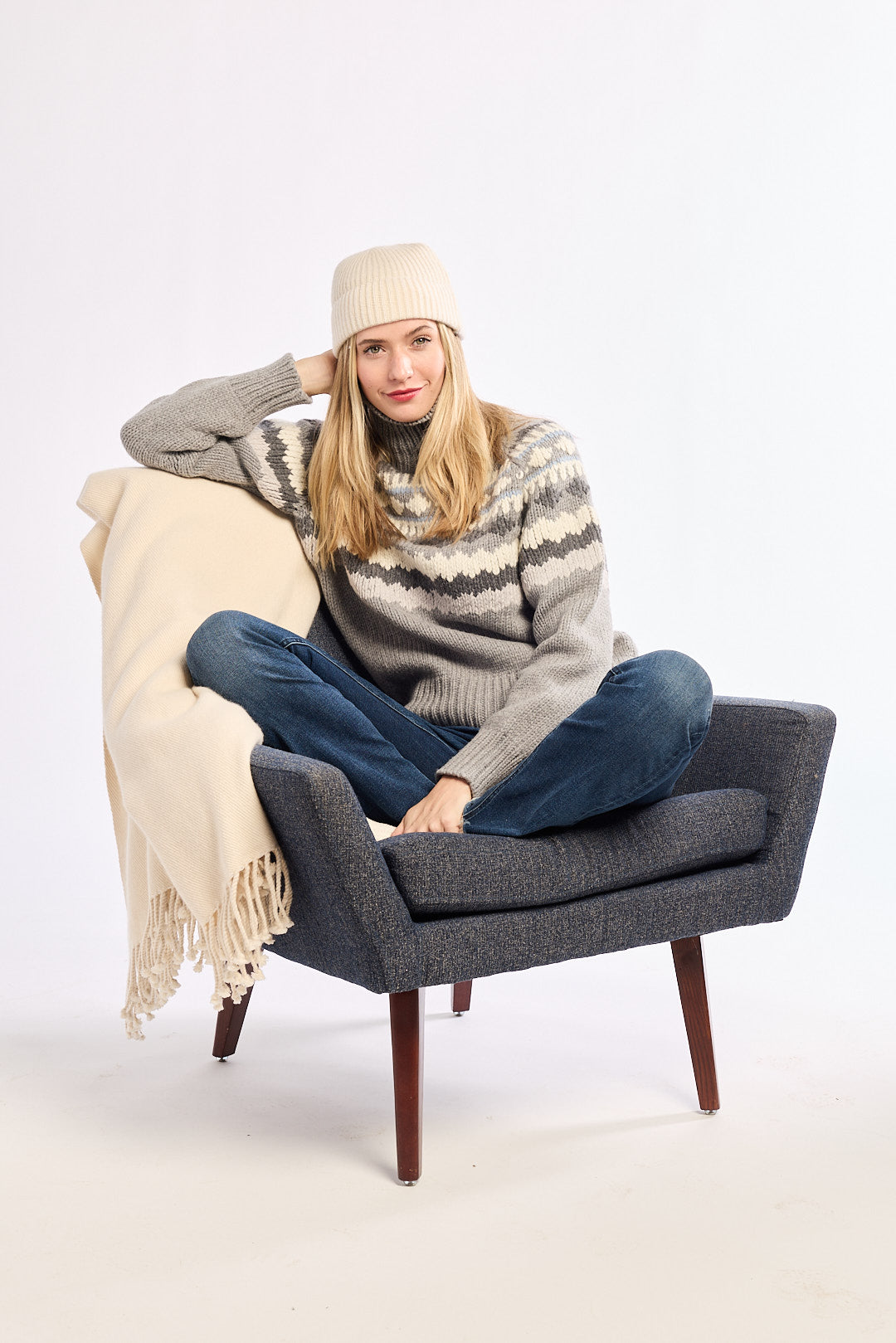 Woman sitting on a gray chair with a beige blanket, wearing a patterned sweater and jeans.