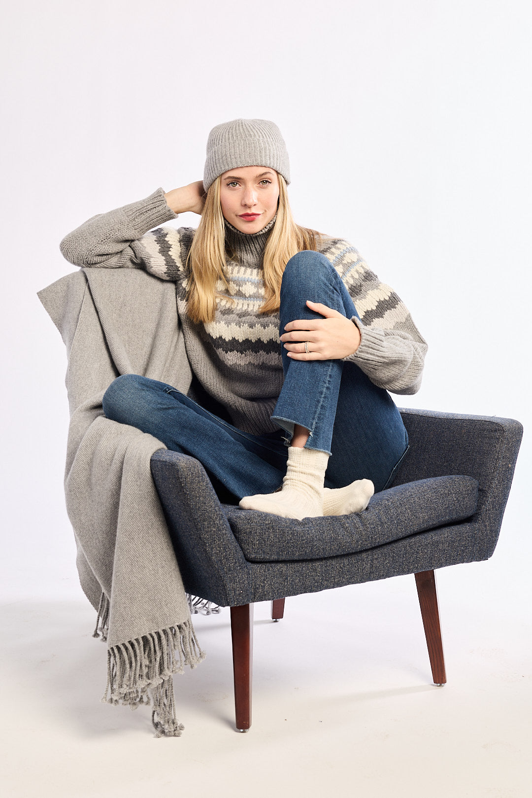 Person sitting on a gray chair wearing a patterned sweater, jeans, and a beanie with a blanket draped over their legs.