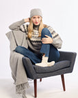 Person sitting on a gray chair wearing a patterned sweater, jeans, and a beanie with a blanket draped over their legs.
