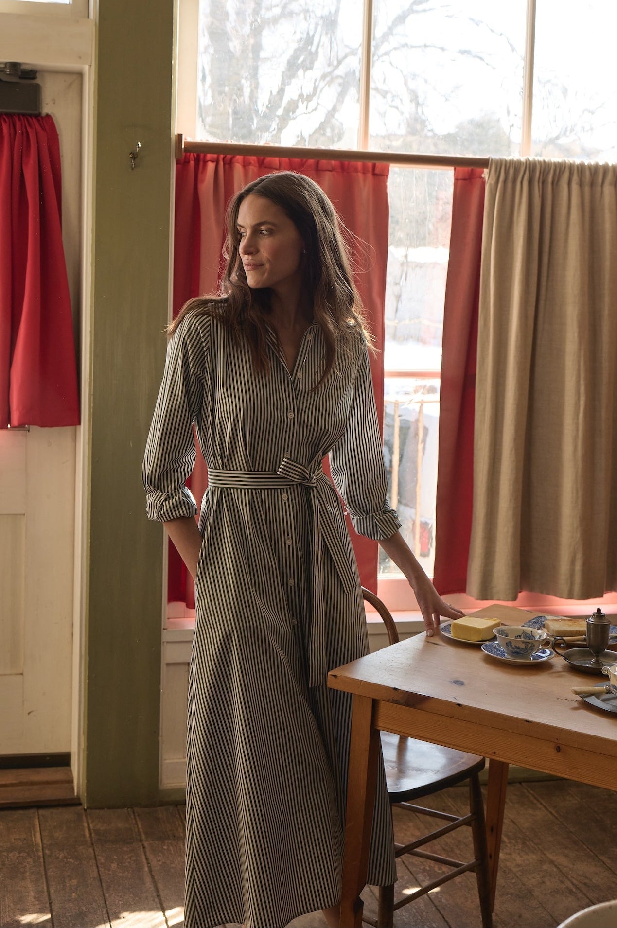 Woman in a striped dress standing in a room with red curtains and a wooden table.