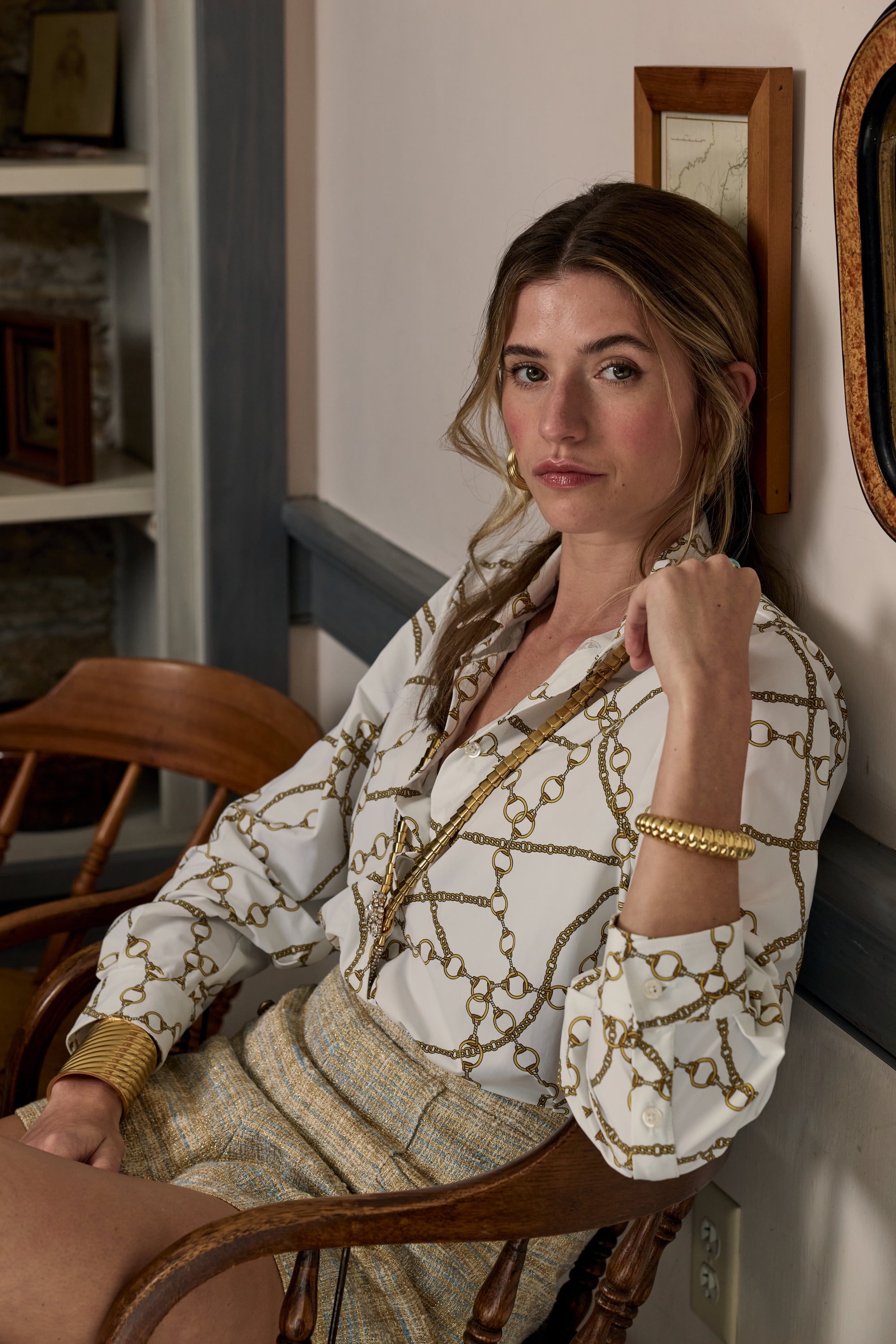 Woman sitting on a wooden chair wearing a patterned blouse in an indoor setting.