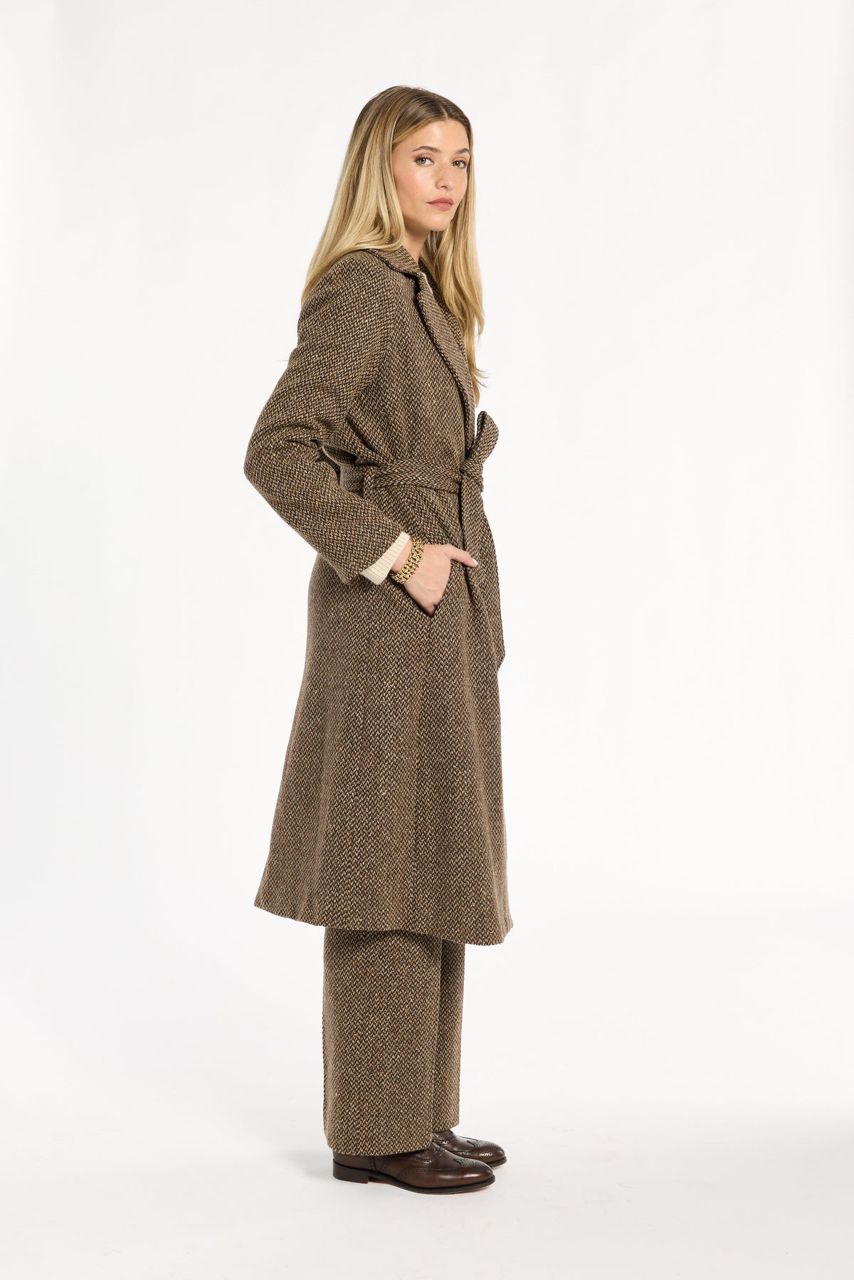A woman modeling the belted Tobacco Tweed Anne Coat with matching trousers, three-quarter side view, photographed against a plain white studio background.