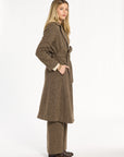 A woman modeling the belted Tobacco Tweed Anne Coat with matching trousers, three-quarter side view, photographed against a plain white studio background.