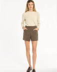 A woman modeling the Tobacco Tweed Archer Shorts with a cream sweater and brown loafers, front view with hands in pockets, photographed against a plain white studio background.