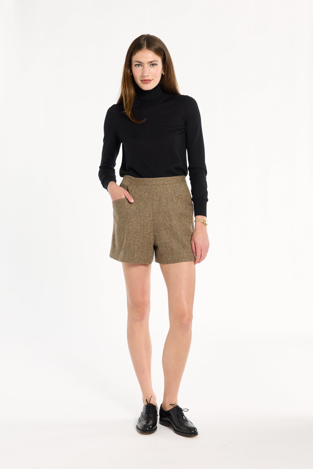A woman modeling the Painter's Fleck Herringbone Archer Shorts with a black turtleneck and black loafers, front view, photographed against a plain white studio background.