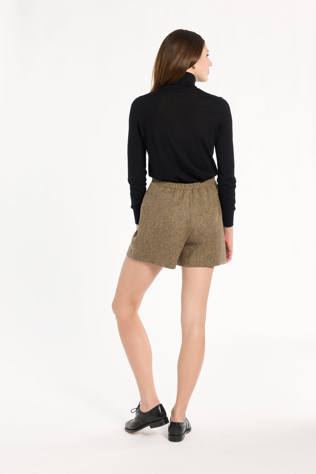 A woman modeling the Painter's Fleck Herringbone Archer Shorts with a black turtleneck and loafers, back view, photographed against a plain white studio background.