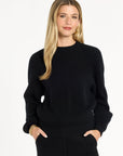 A woman modeling the black Baby Cashmere Ribbed Crewneck with black trousers, front view with hands in pockets, photographed against a plain white studio background.