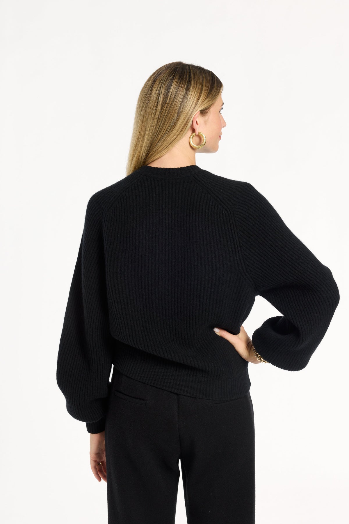 A woman modeling the black Baby Cashmere Ribbed Crewneck with black trousers, back view, photographed against a plain white studio background.