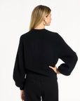 A woman modeling the black Baby Cashmere Ribbed Crewneck with black trousers, back view, photographed against a plain white studio background.
