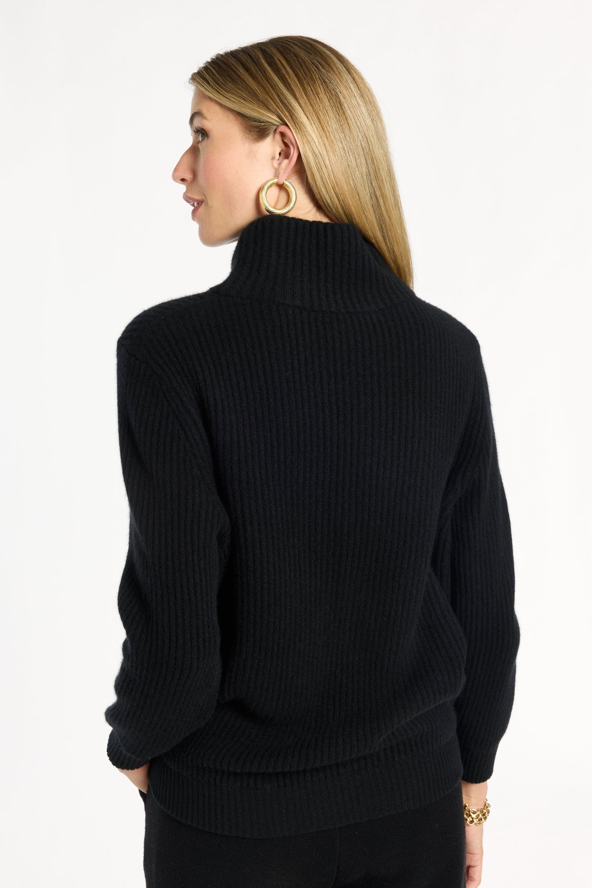 A woman modeling the black Baby Cashmere Ribbed Turtleneck with black trousers, back view, photographed against a plain white studio background.