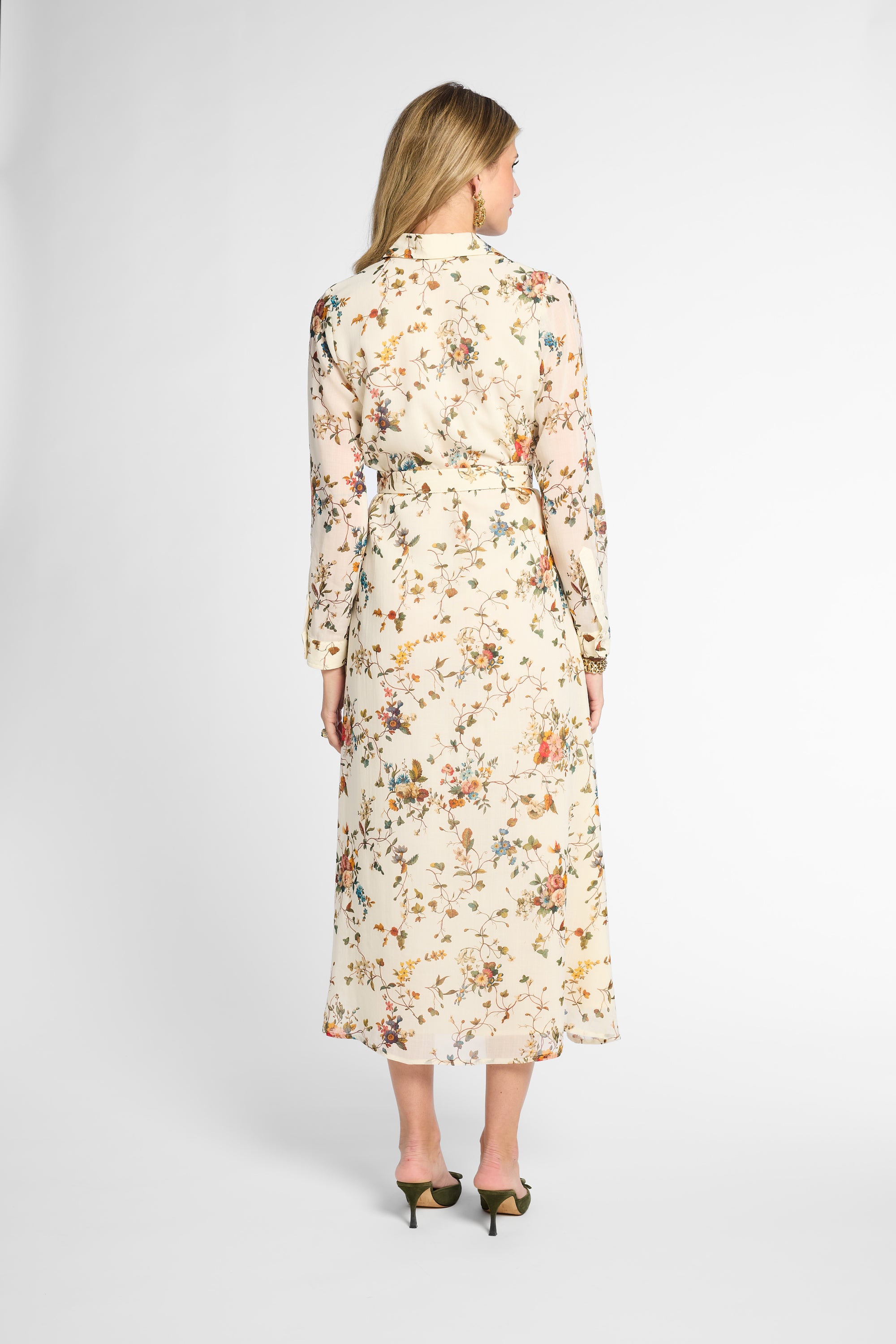 A woman viewed from the back wearing a long-sleeve midi dress with a cream base and an all-over multicolor floral print, paired with black pointed-toe heels against a light gray studio background.