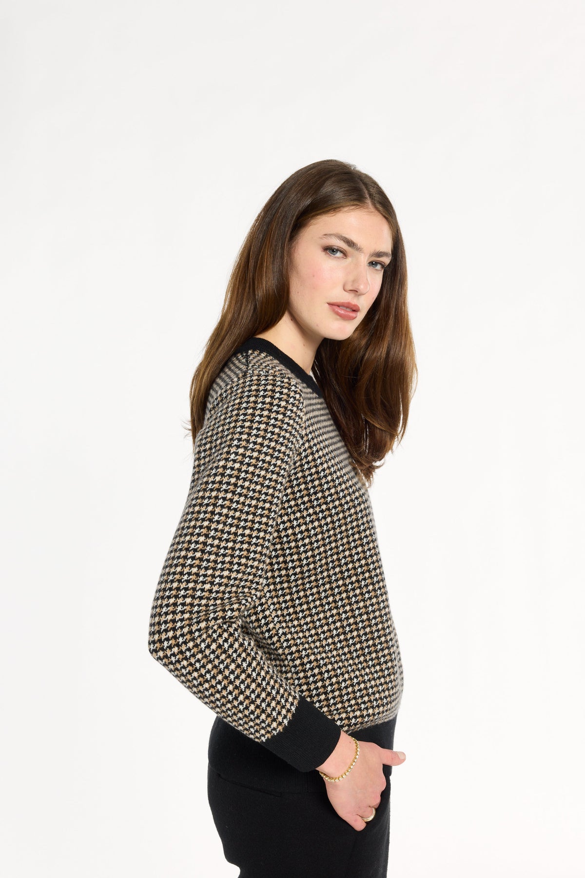 A woman modeling the Black Tri-Color Check Classic Cashmere Crew with black trousers, side view with one hand in a pocket, photographed against a plain white studio background.