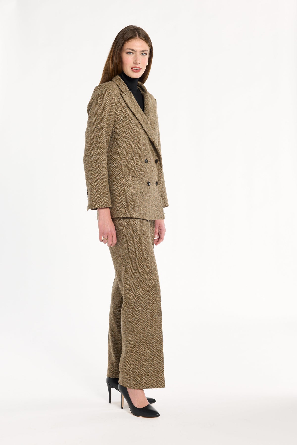 A woman modeling the Painter's Fleck Herringbone Cynthia Blazer with matching trousers, side view, photographed against a plain white studio background.