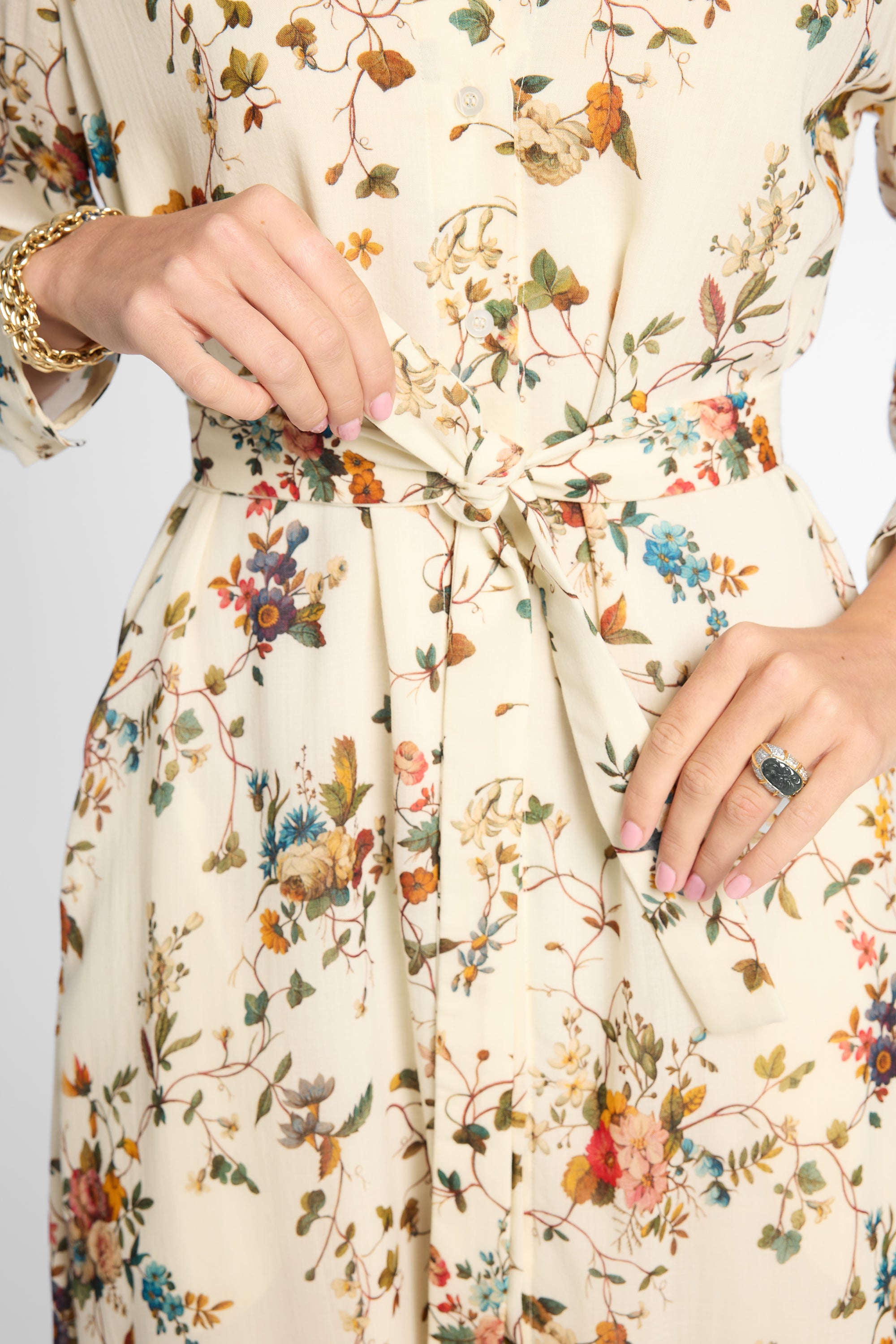 Close-up of a person wearing a cream-colored long-sleeve dress with a multicolor floral and bird print, featuring a tie waist belt and pockets.