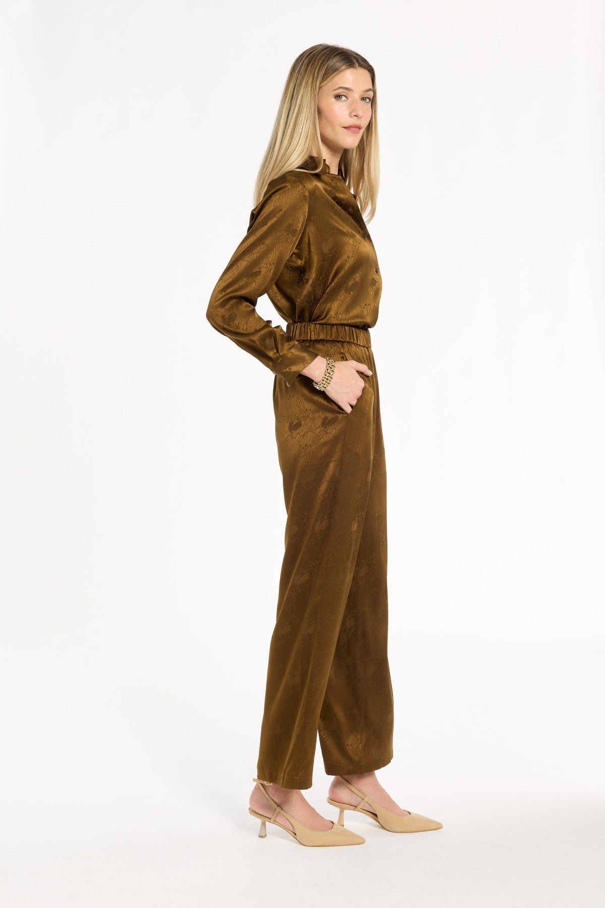 A woman modeling the Golden Umber Diane Pants with the coordinating Eleanor Shirt and cream heels, side view, photographed against a plain white studio background.