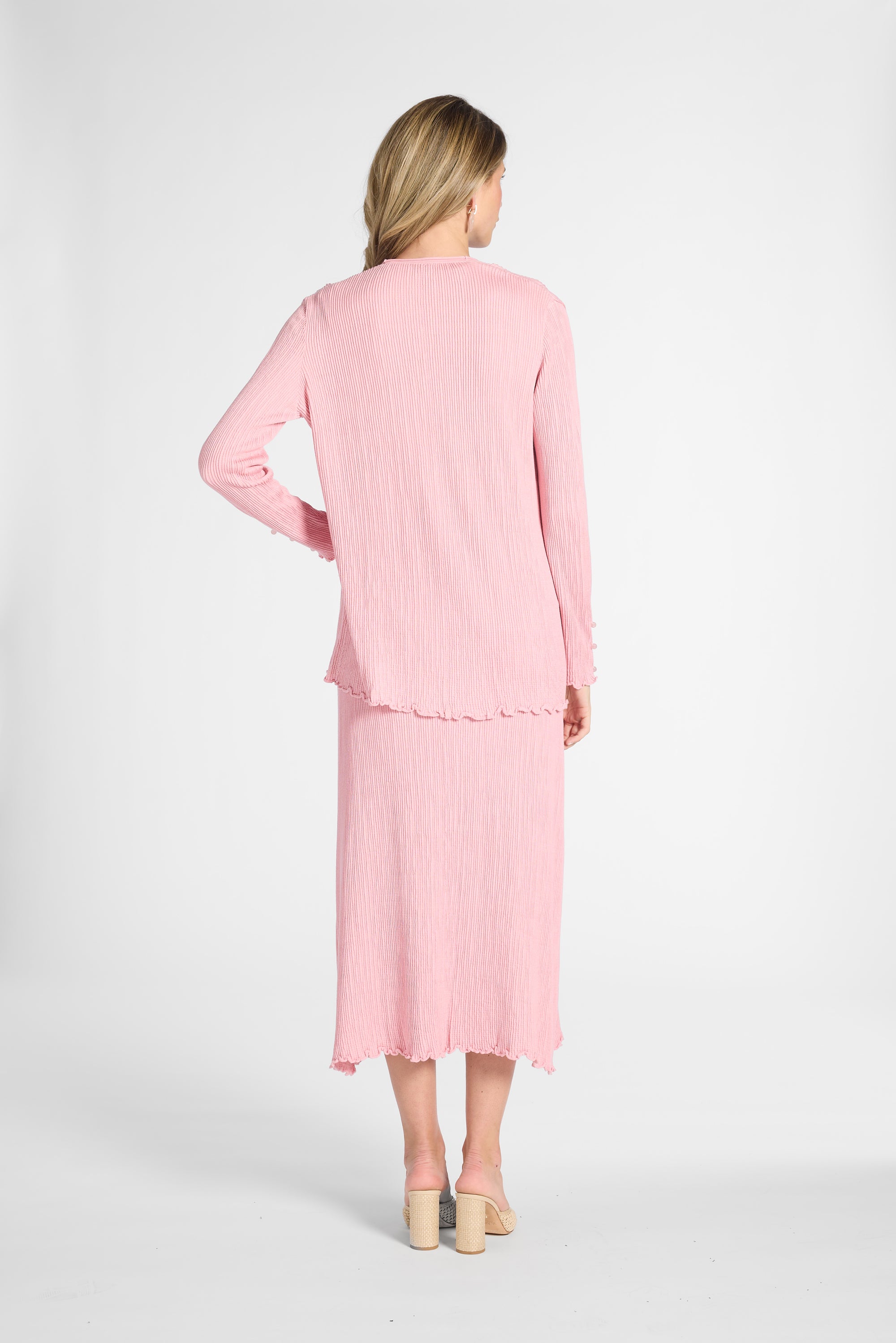 Woman standing to the back modeling a pink cardigan and the matching pleated skirt on a white backdrop