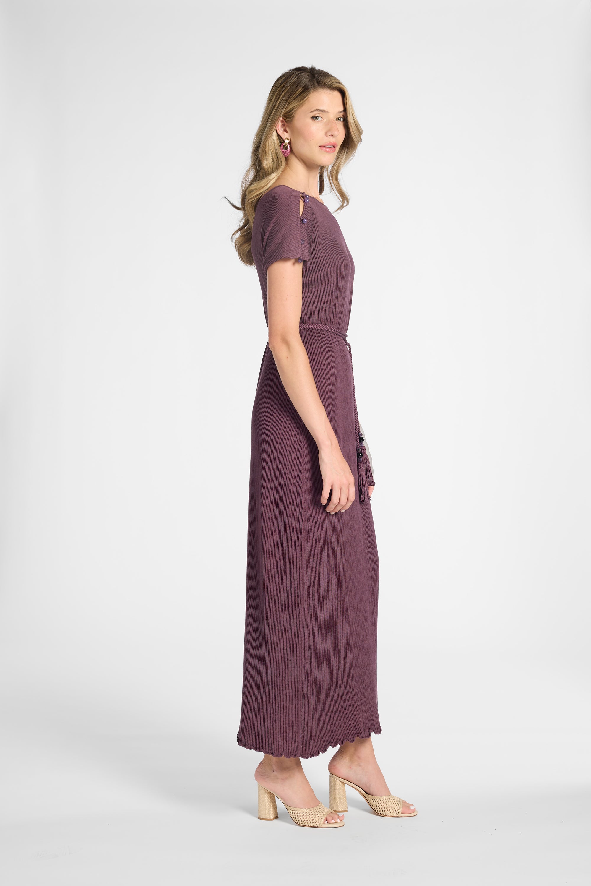 A woman modeling a short-sleeved, floor-length purple dress with a tie waist, shown in profile against a light gray studio background.