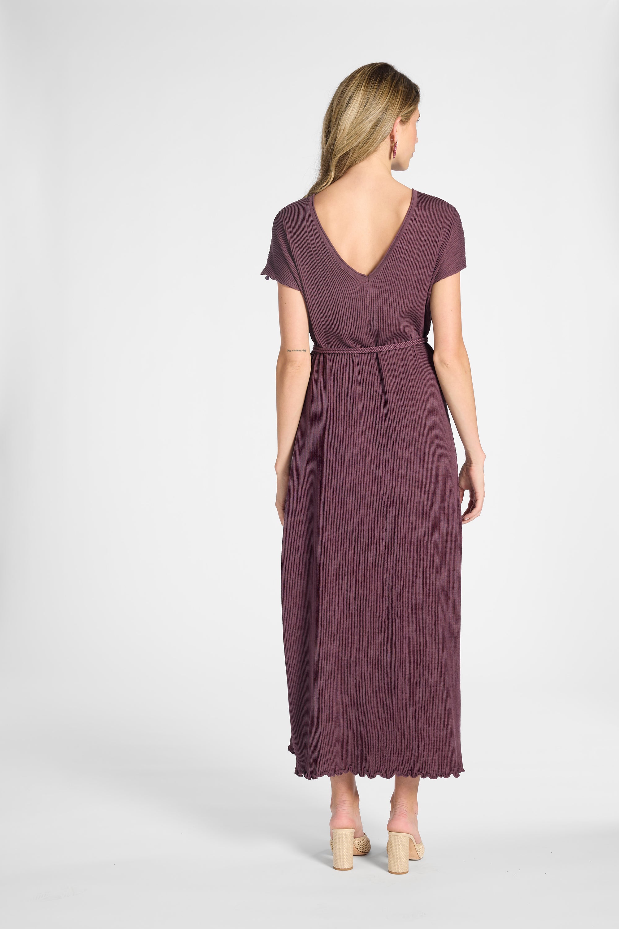 A woman viewed from the back wearing a short-sleeve, V-neck maxi dress in a muted plum-brown color, paired with beige high-heeled sandals, against a light gray studio background.