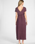 A woman viewed from the back wearing a short-sleeve, V-neck maxi dress in a muted plum-brown color, paired with beige high-heeled sandals, against a light gray studio background.