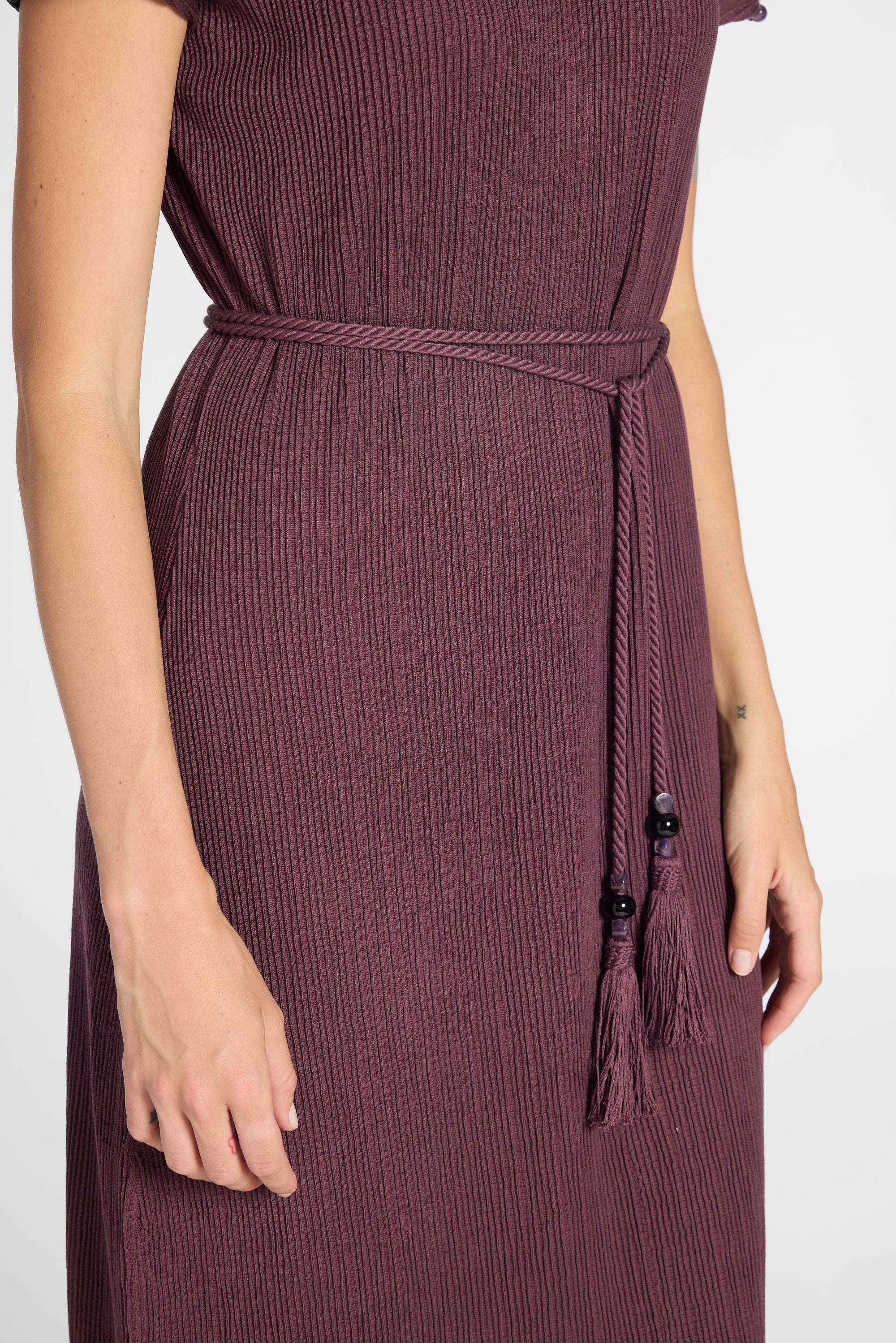 A close-up of a woman wearing a ribbed purple dress with a matching tie belt and tassels, shown against a plain light background.
