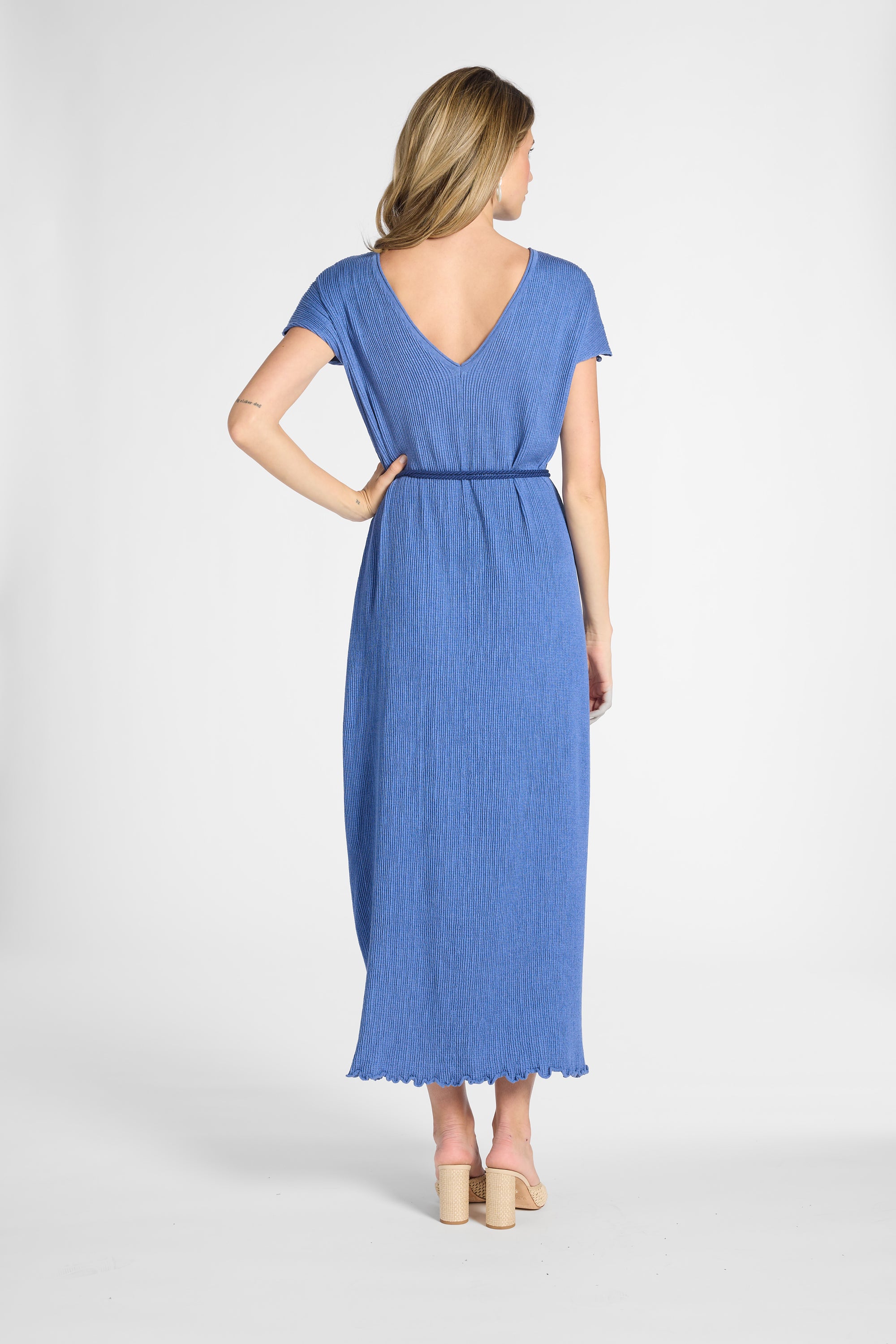 A woman modeling a blue midi dress from the back, featuring a V-neckline, short sleeves, a matching fabric tie belt at the waist, and a scalloped hem, set against a plain light gray studio background.