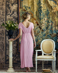 A woman stands with her back to the camera wearing a light pink midi dress, posing in front of a large vintage-style tapestry. To her left is a cream pedestal with a flower pot, and to her right is a white upholstered armchair.