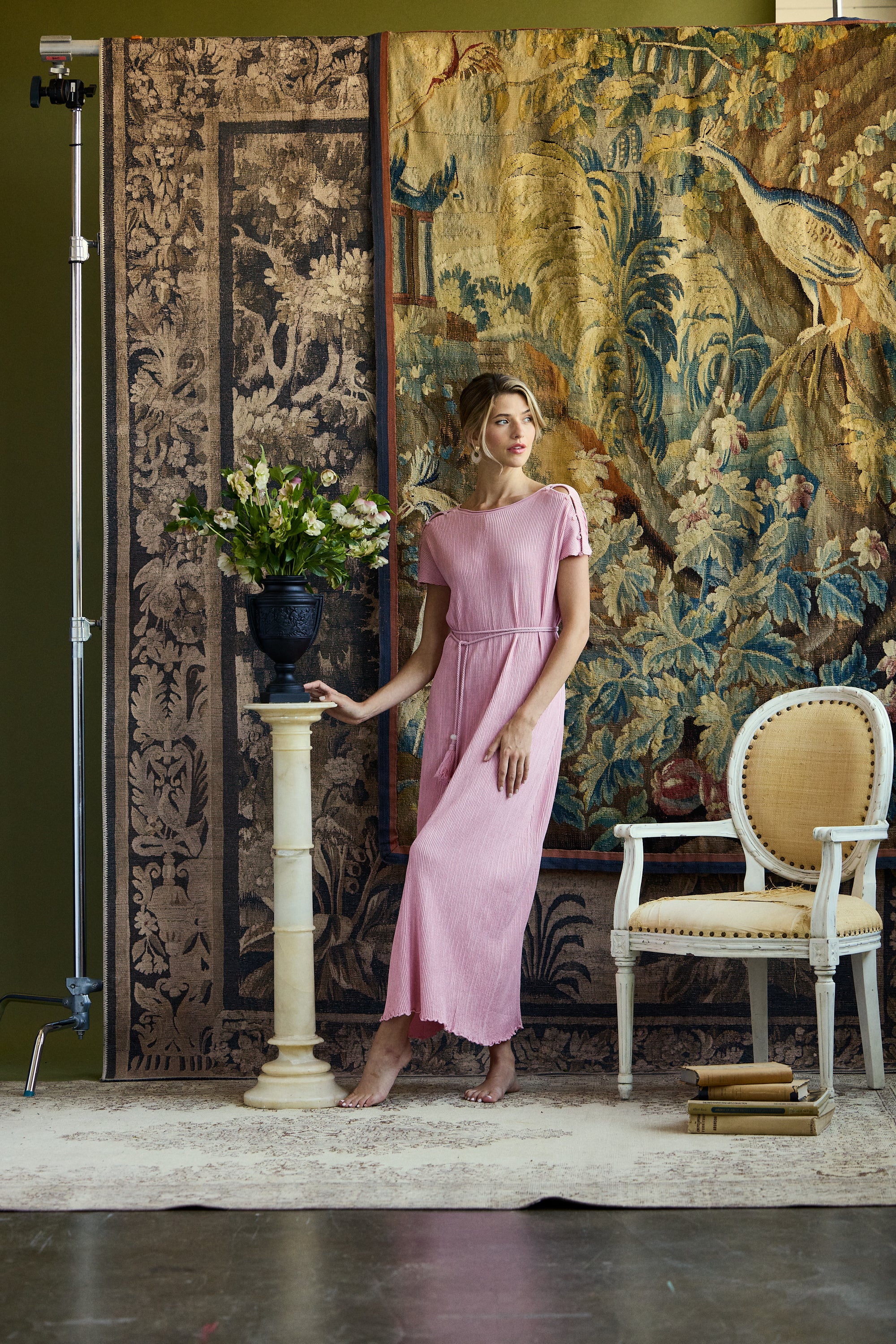 A woman models a light pink midi dress with short sleeves and a gathered waist, standing in a styled studio setting with a large tapestry backdrop, a pedestal with flowers, and a white armchair.