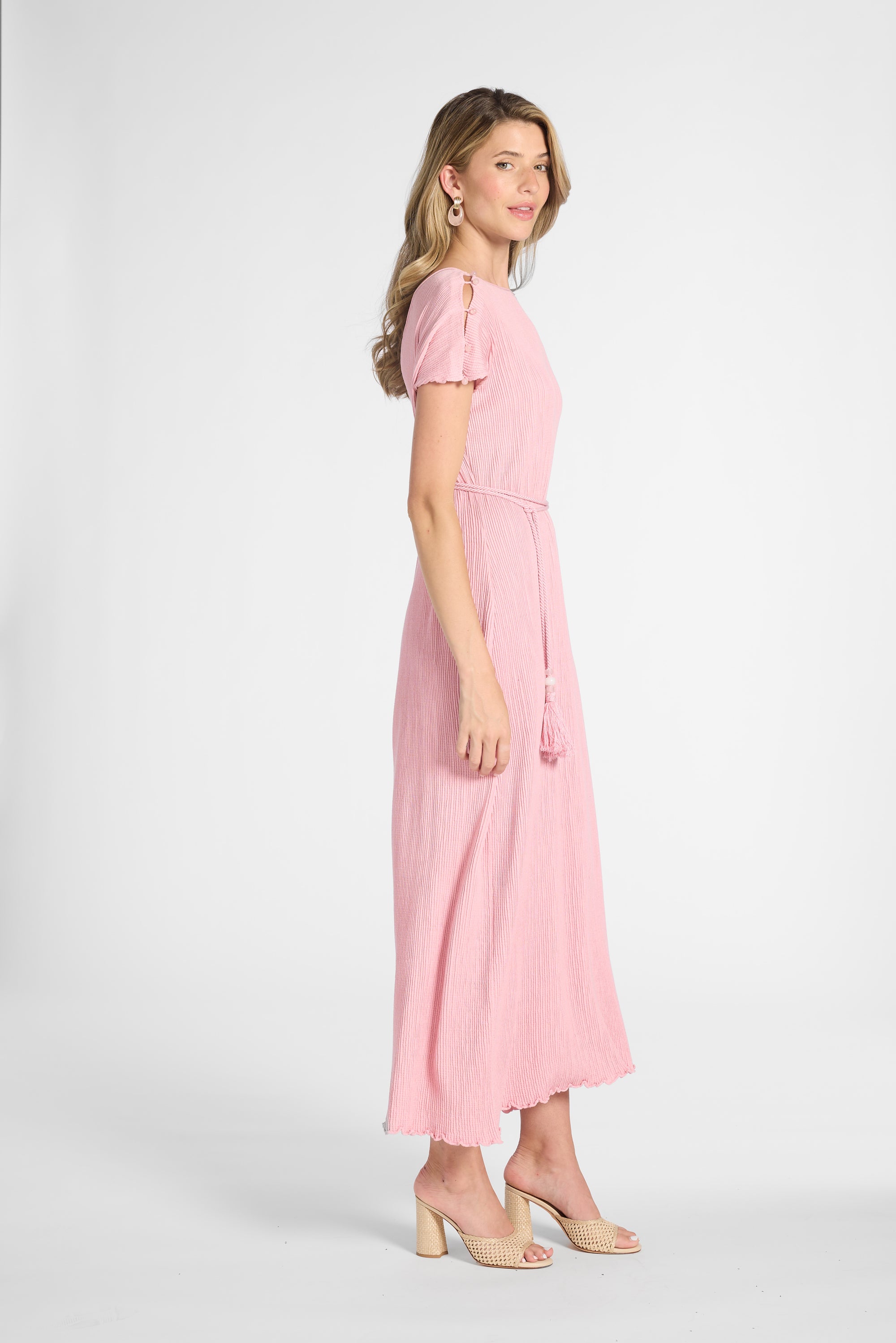 A woman modeling a light pink midi dress with short puff sleeves, a matching tie belt, and tassel details, paired with beige heeled sandals against a light gray studio background.