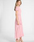 A woman modeling a light pink midi dress with short puff sleeves, a matching tie belt, and tassel details, paired with beige heeled sandals against a light gray studio background.