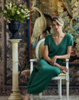 A woman sits barefoot on a white wooden chair in a decorated room, wearing a dark green V-neck maxi dress. 