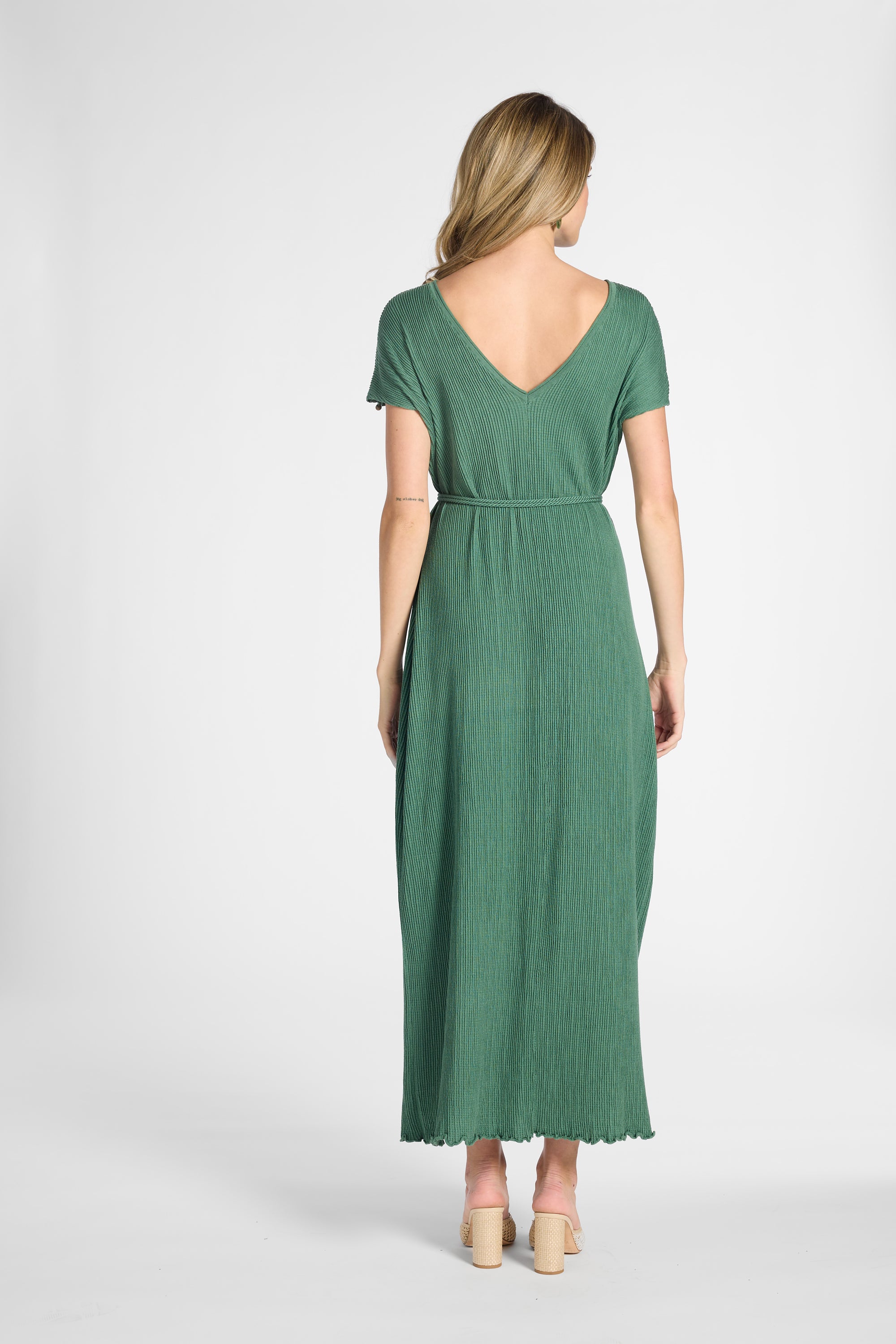 A woman viewed from the back wearing a green short-sleeve maxi dress with a V-neckline and tie waist, paired with beige heels, against a light gray studio background.