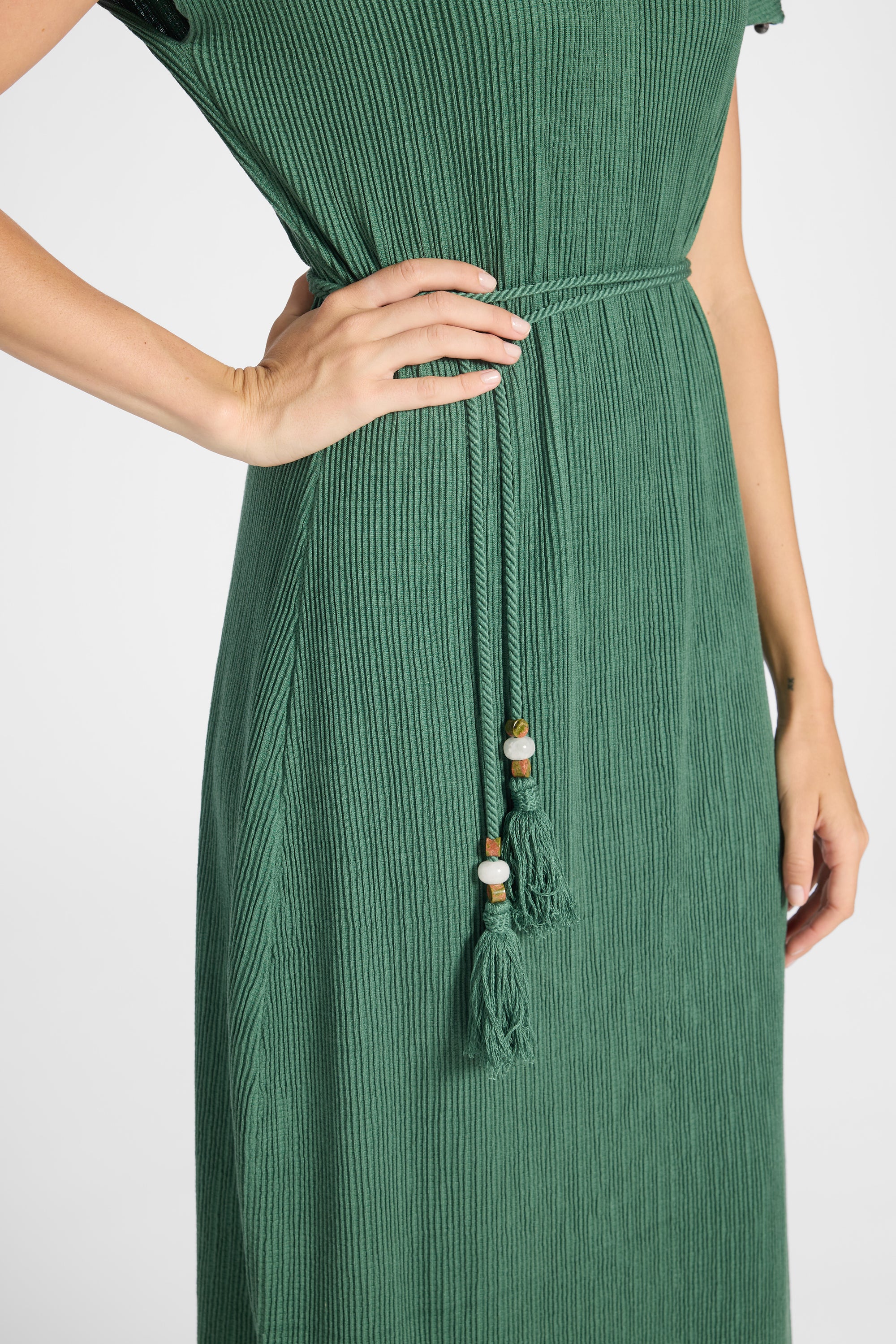 A close-up of a woman wearing a green pleated maxi dress with a tie-waist belt and tassel detail, shown against a plain light background.